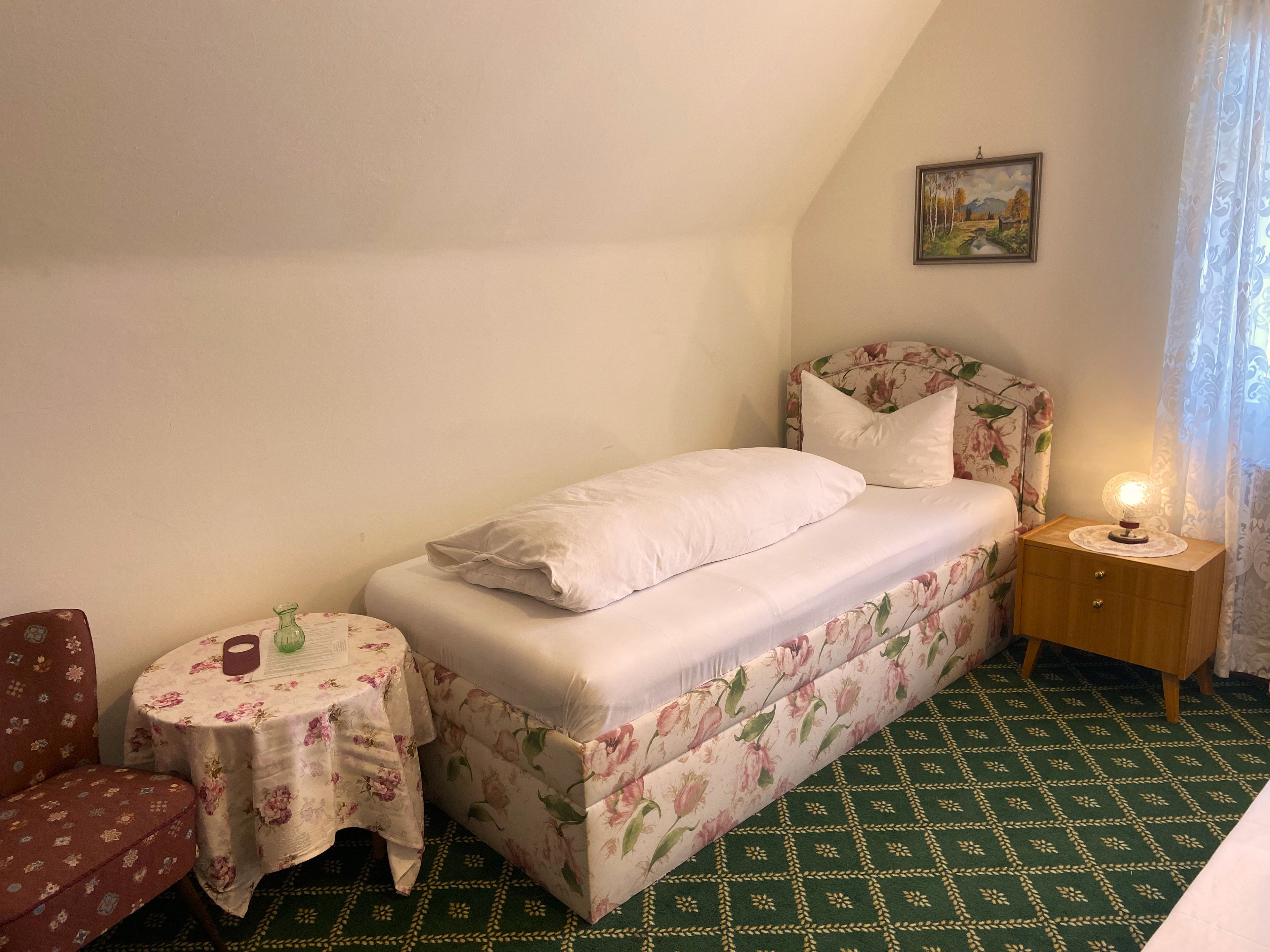 Une petite pièce agréable, avec un lit, une petite table ronde, une chaise et une table de chevet avec une lampe.