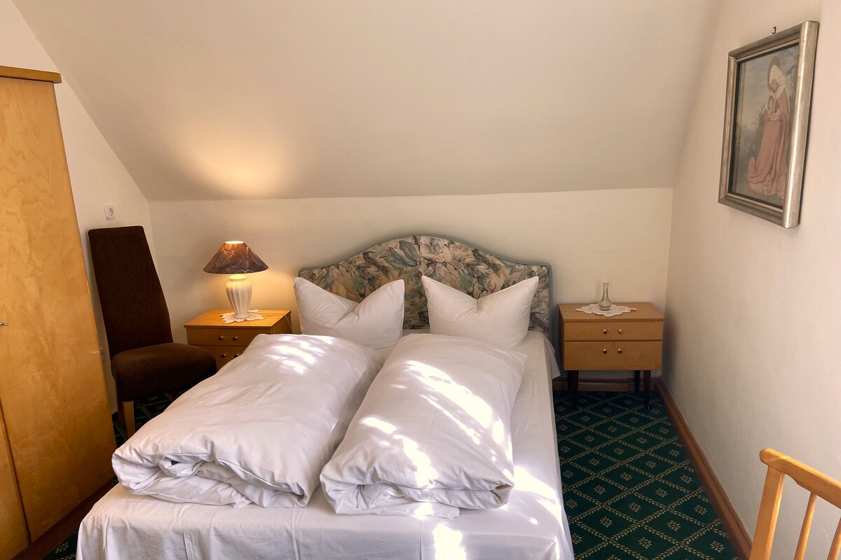 Habitación de hotel con cama, mesita de noche con lámpara, una silla, armario de madera y alfombra estampada.