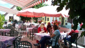 Een terras met tafels en stoelen onder gestreepte luifels, omringd door groen en gebouwen.