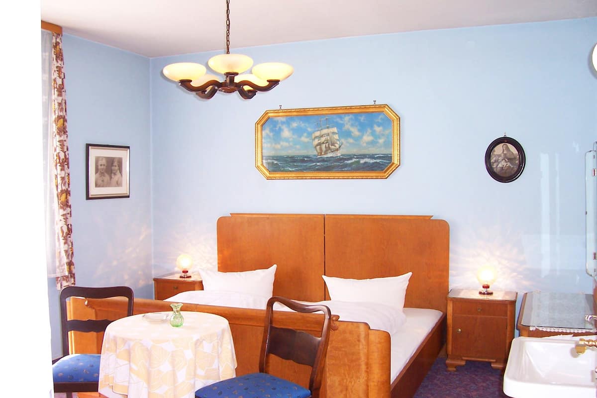 Una habitación de hotel con una cama, dos mesitas de noche, una silla, una mesita pequeña con una lámpara, un cuadro de un barco en la pared y un espejo.