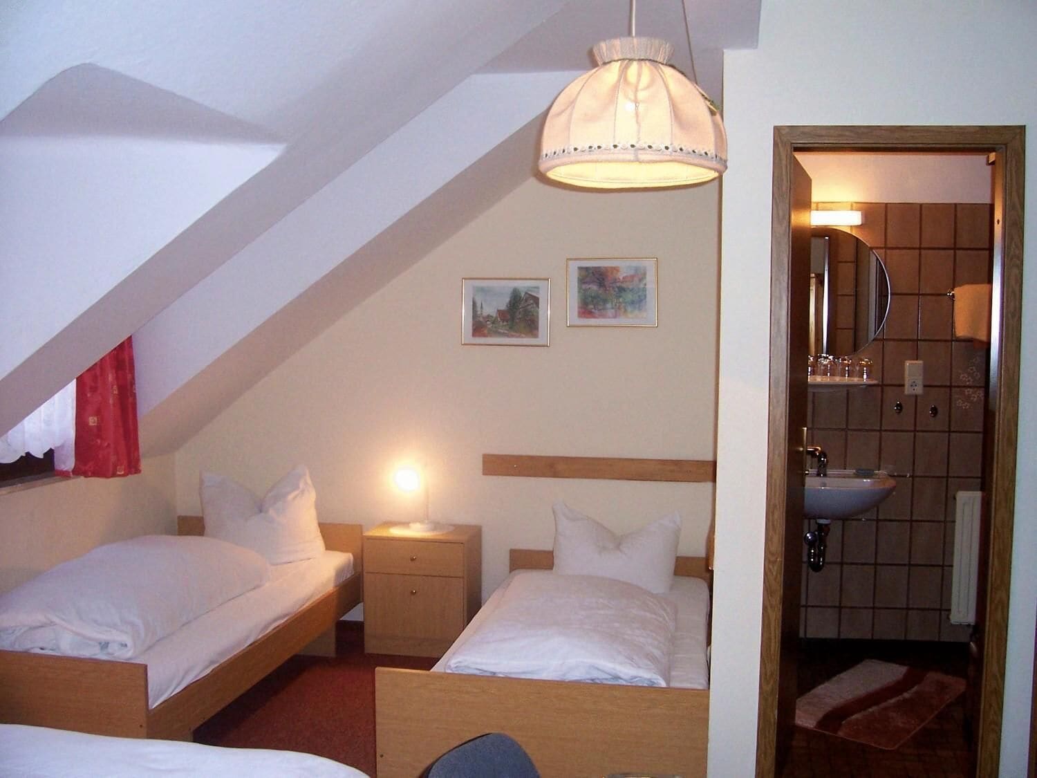 A small, cozy room with two single beds, a nightstand, a lamp, and a bathroom visible through an open door.