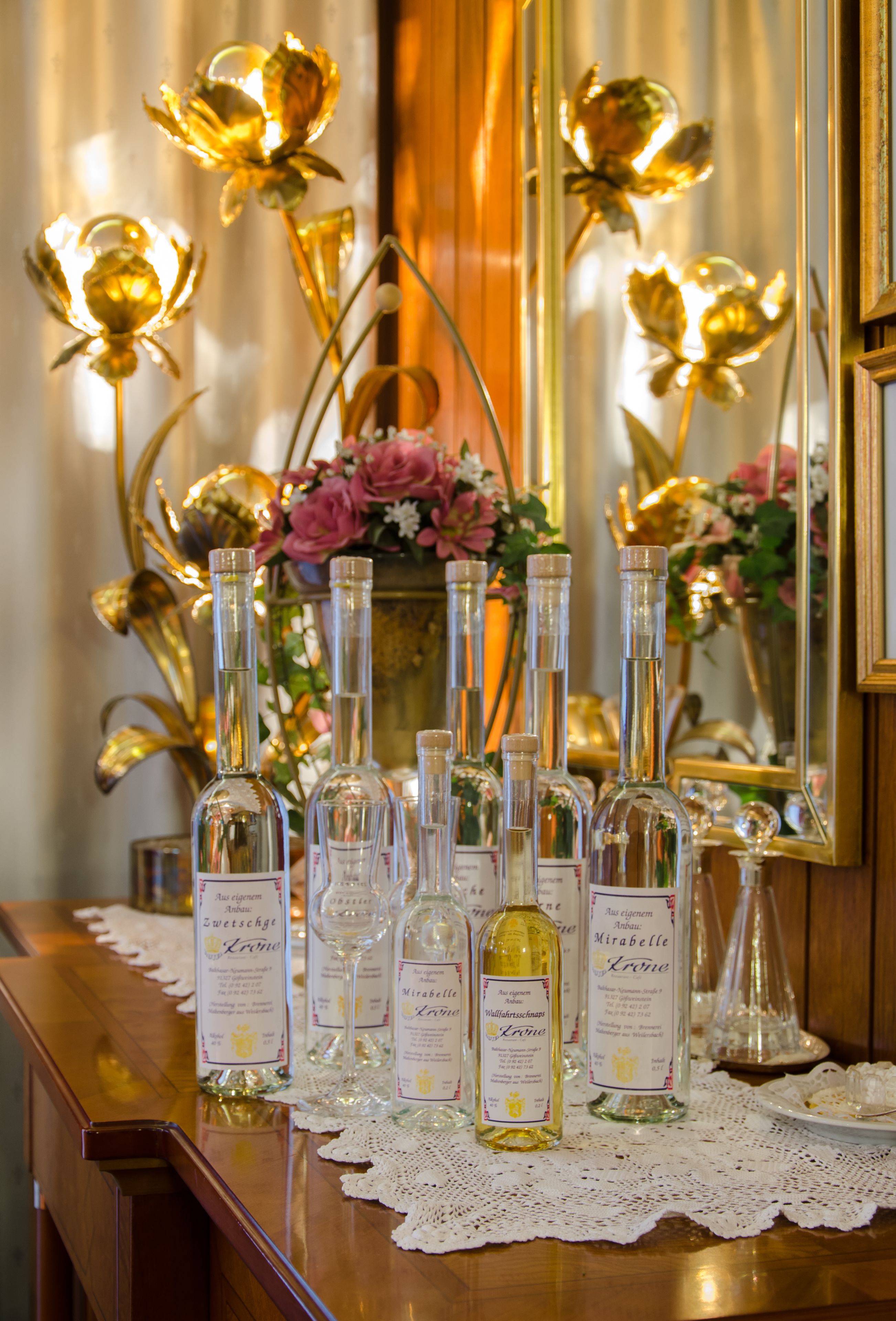 Un espace bar avec des bouteilles d’alcool et des verres disposés sur un comptoir en bois, un miroir et des fleurs décoratives en arrière-plan.
