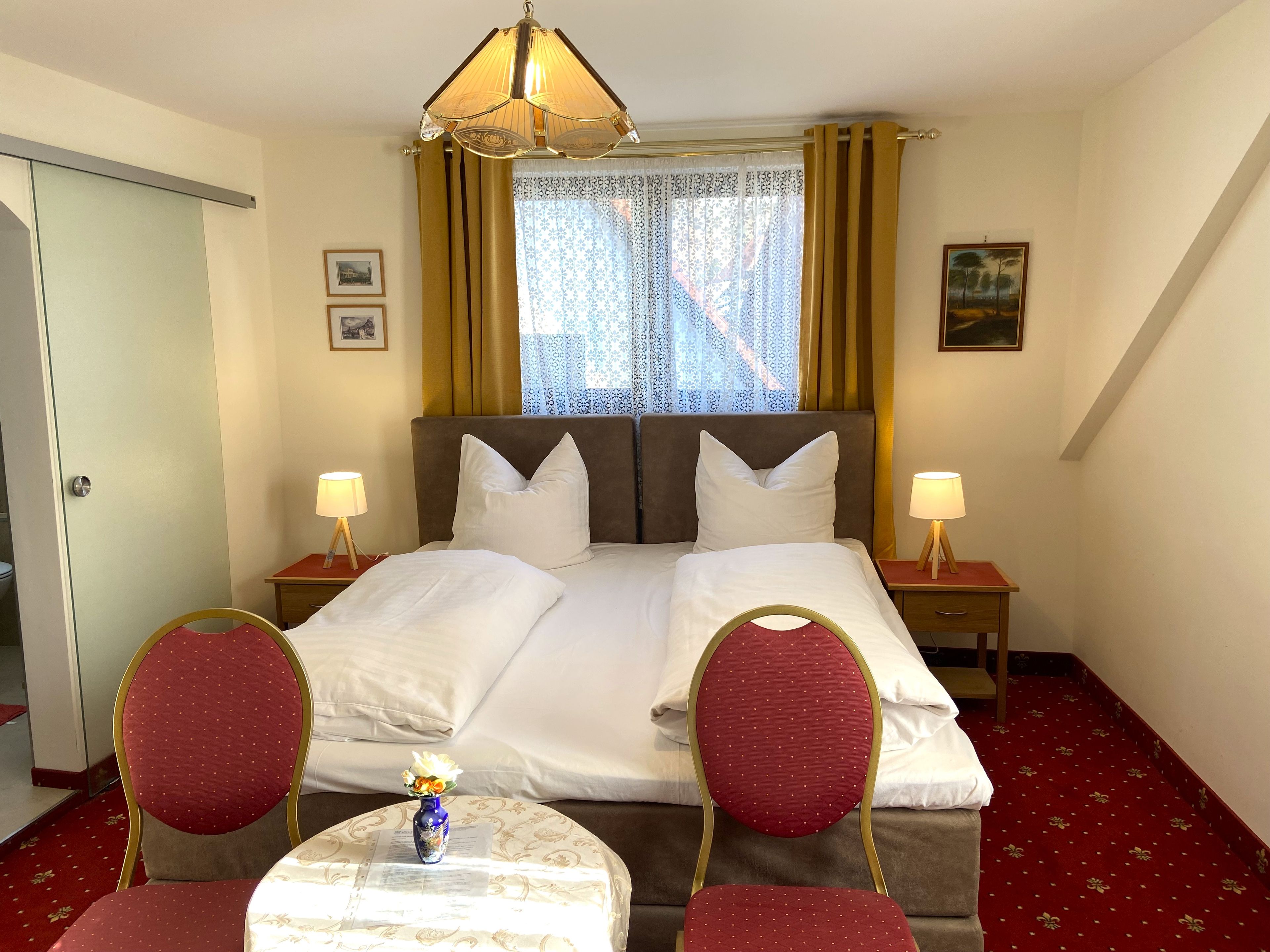 Una habitación de hotel con una cama, dos mesitas de noche con lámparas, una silla, una pequeña mesa con un florero y una ventana con cortinas.