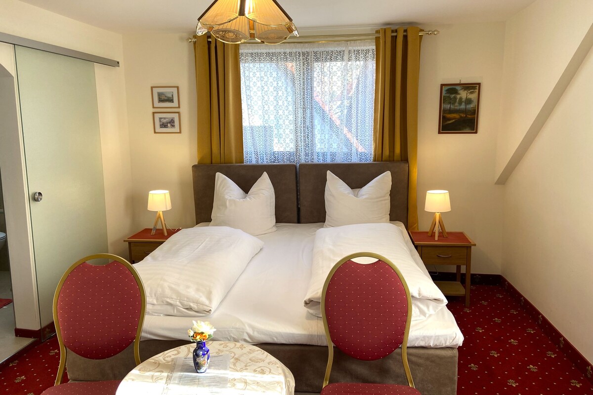 Una habitación de hotel con una cama, dos mesitas de noche con lámparas, una silla, una pequeña mesa con un florero y una ventana con cortinas.