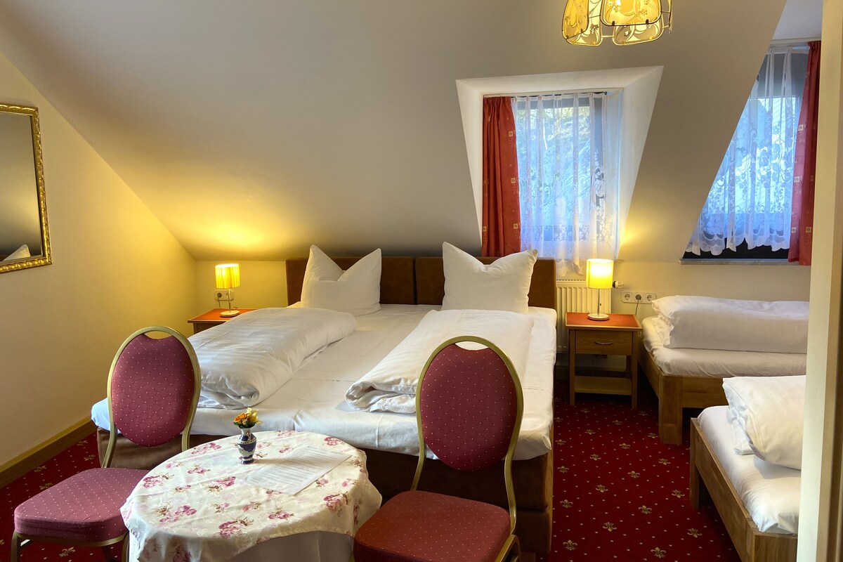 Una habitación de hotel con cama, dos mesitas de noche, una mesa con sillas, un espejo y un candelabro.