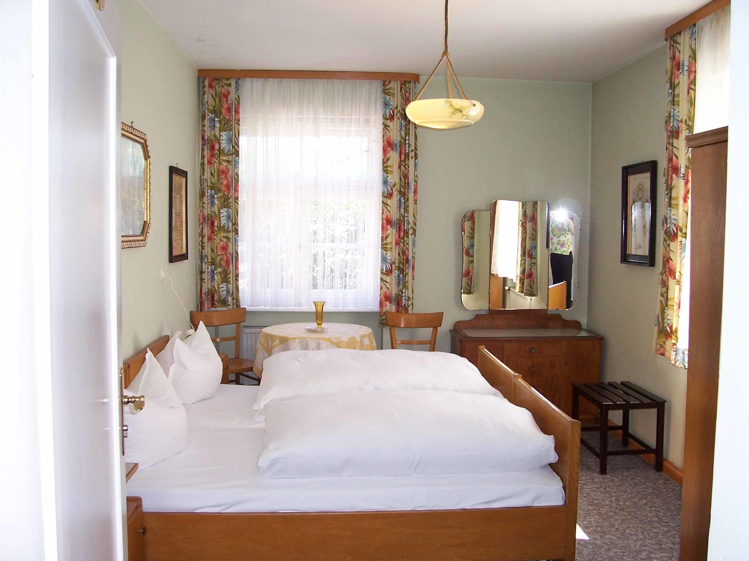 Ein Hotelzimmer mit einem großen Bett, einem kleinen Tisch, einem Stuhl, einem Schreibtisch, einem Spiegel und blumigen Vorhängen.