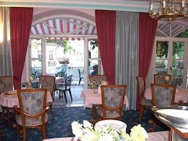 Uma sala de jantar com mesas e cadeiras, um lustre e grandes janelas com cortinas.