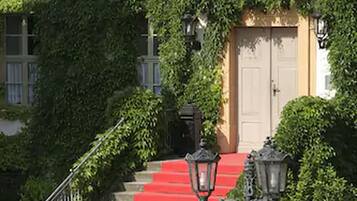 Un edificio con una escalera alfombrada de color rojo que conduce a una puerta blanca, rodeado de abundante vegetación.