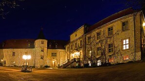 Un edificio histórico de piedra con una torre, ventanas iluminadas y un patio empedrado.