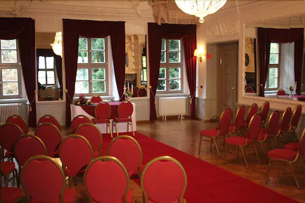 Un salón elegante con sillas rojas dispuestas en filas, una mesa con mantel blanco y amplias ventanas con cortinas.
