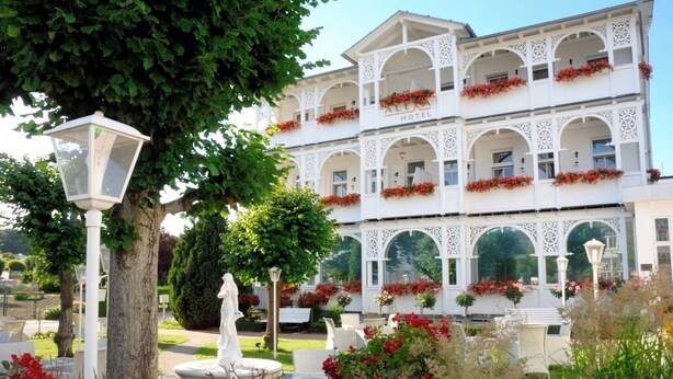 A multi-story hotel with white architecture, balconies with red flowers, and a garden with a statue and lamp posts. - Alexa Hotel (Göhren)
