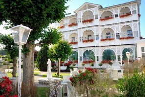 Un hôtel à plusieurs étages, doté d’une architecture blanche, de balcons ornés de fleurs rouges et d’un jardin agrémenté d’une statue et de lampadaires.