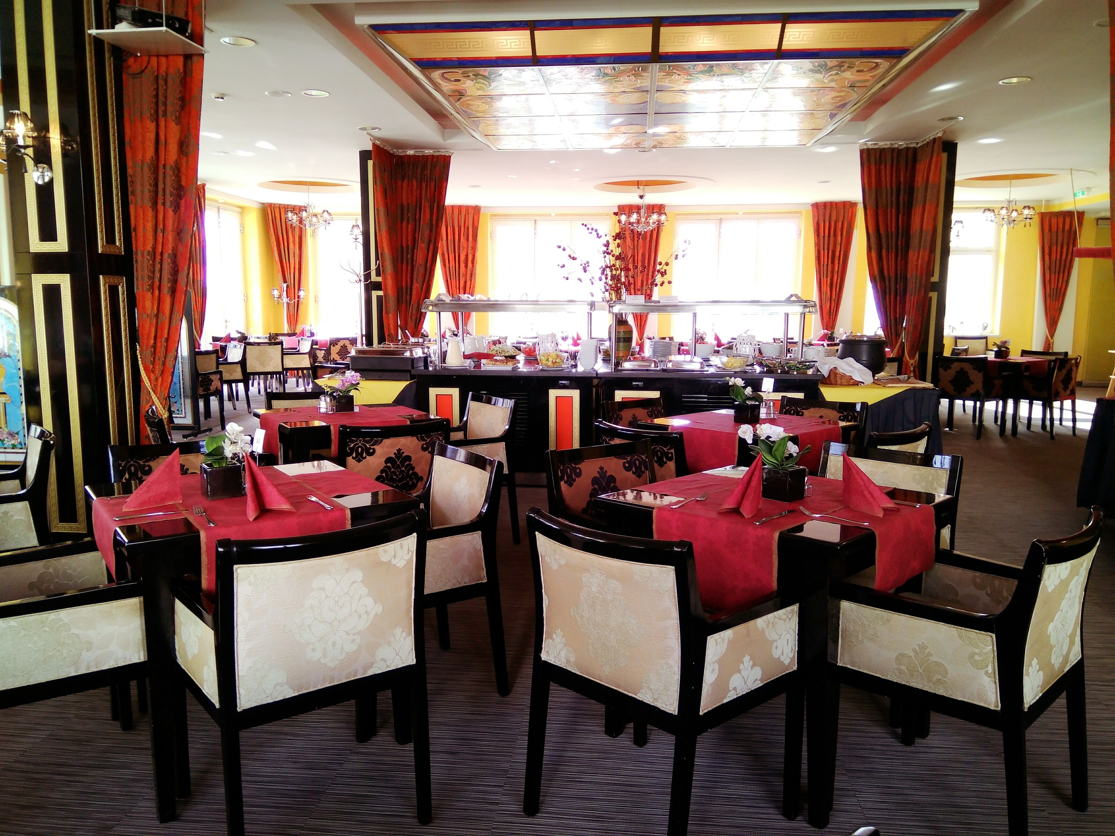 Un restaurante con varias mesas y sillas, una zona de bar central y detalles decorativos como cortinas y arreglos florales.