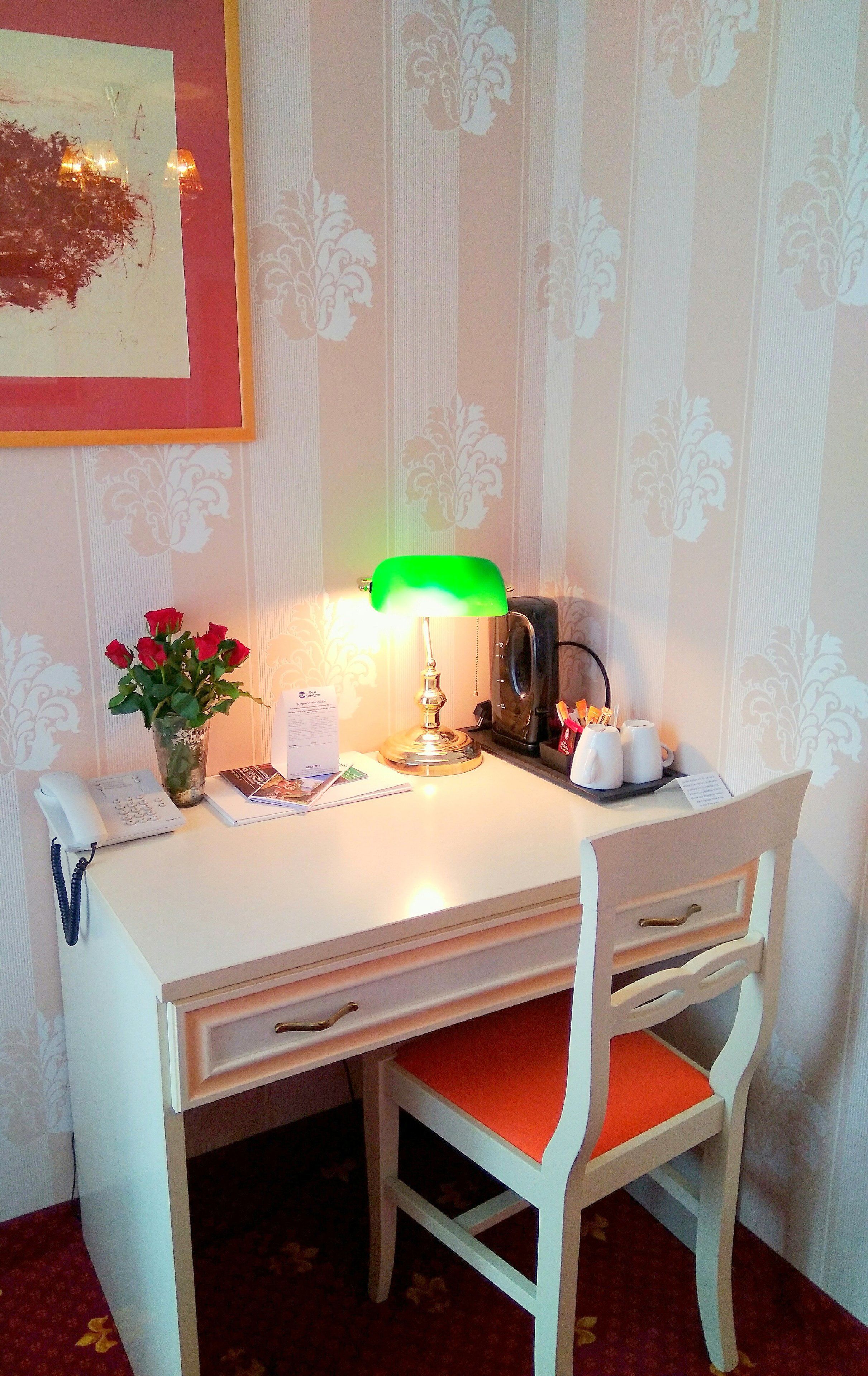 Un petit bureau blanc, sur lequel se trouvent une lampe de bureau verte, un téléphone, un vase de roses rouges et une machine à café noire.