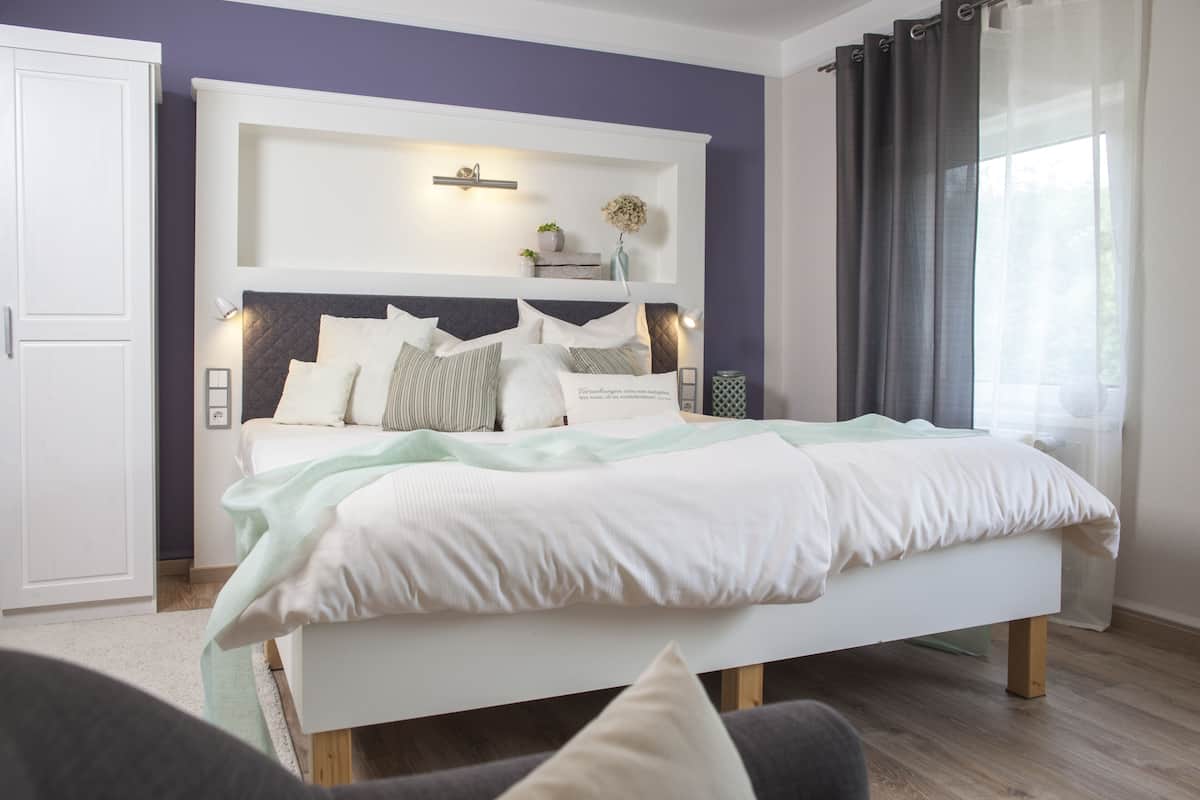 A modern bedroom with a white bed, a headboard with shelves, and a window with curtains.