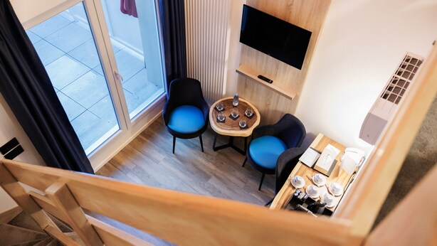 Ein modernes Hotelzimmer mit einer Holztreppe, einem an der Wand befestigten Fernseher, zwei blauen Stühlen um einem kleinen Holztisch und einer Kaffeestation.