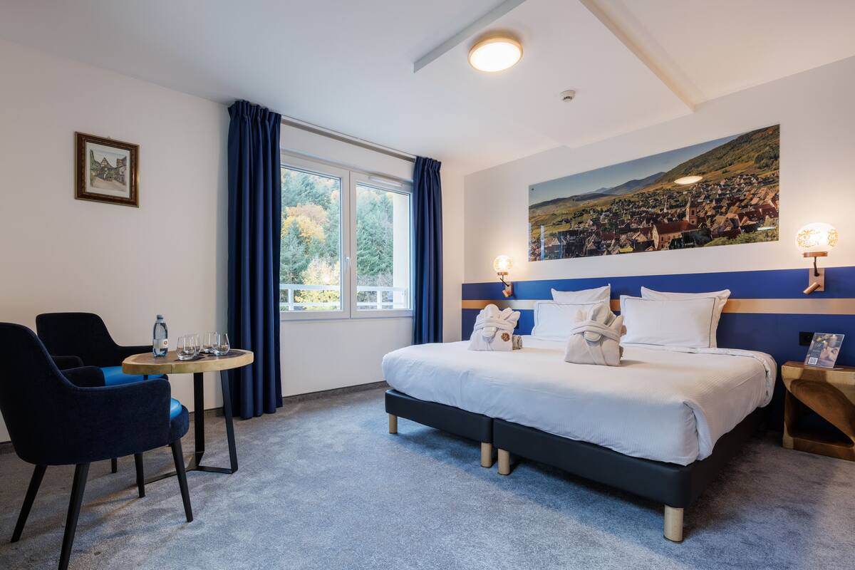 Una habitación de hotel con cama, escritorio, silla, ventana con cortinas y un cuadro en la pared.
