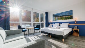 Una habitación de hotel moderna con una cama grande, un sofá, dos sillones, una mesita y vistas al exterior a través de amplios ventanales.