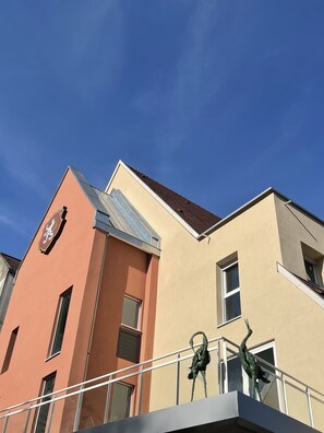 A building with a beige facade, a brown section with a crest, and a balcony with a metal sculpture. - Hotel SPA Husseren Collections - Proche Colmar - Eguisheim (Husseren-les-Chateaux)