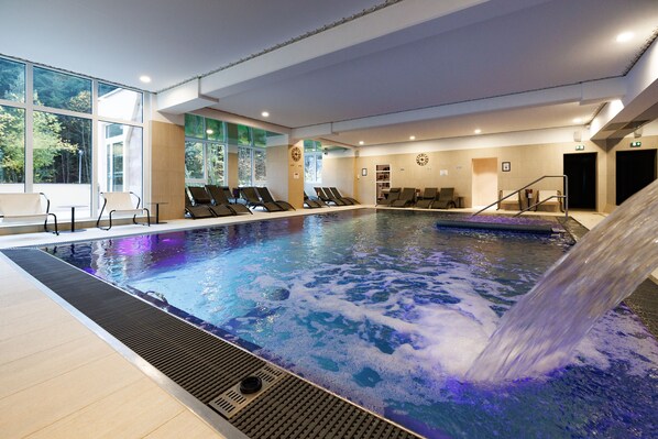 A large indoor swimming pool with a waterfall feature, surrounded by lounge chairs and large windows. - Hotel SPA Husseren Collections - Proche Colmar - Eguisheim (Husseren-les-Chateaux)