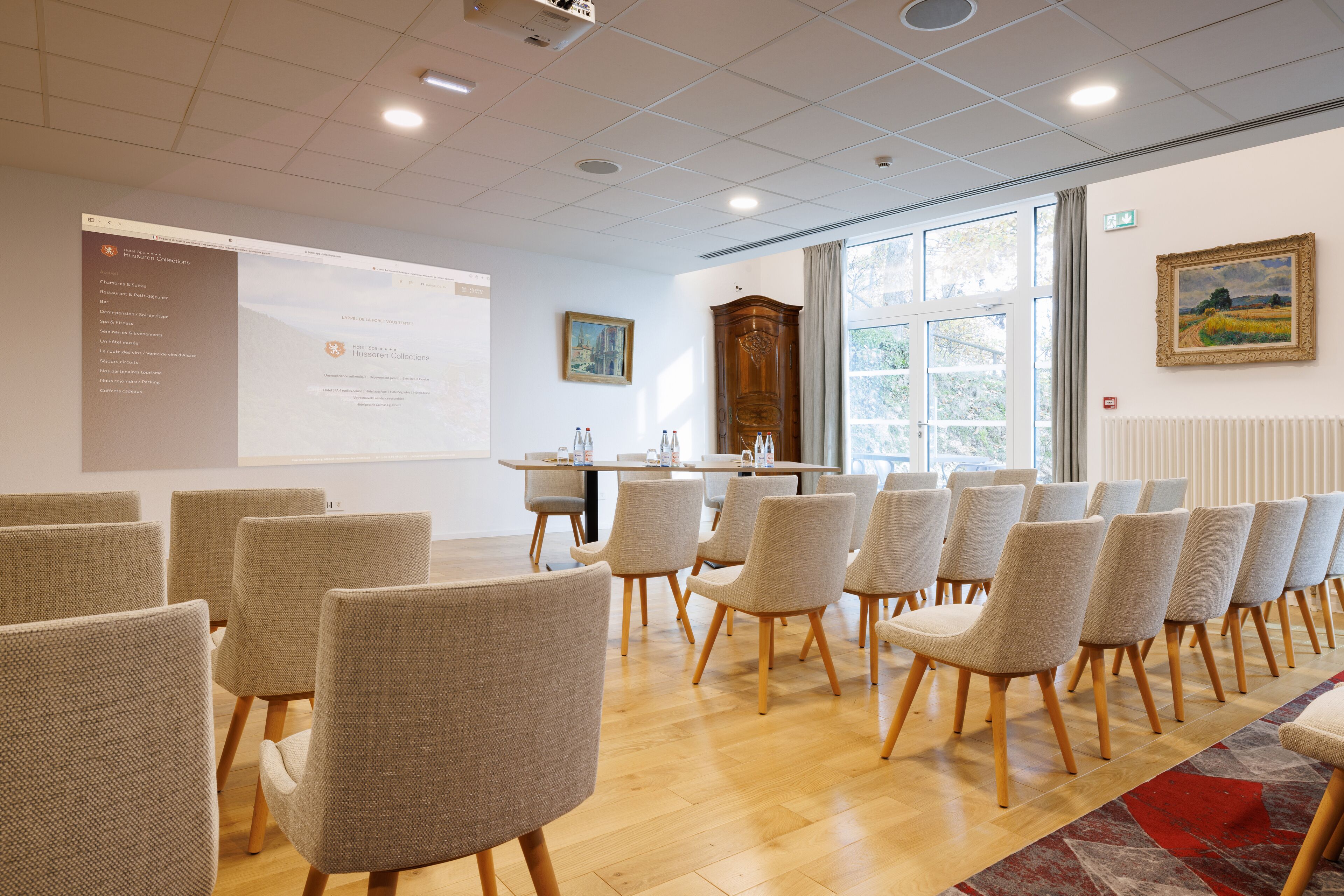 A conference room with rows of chairs, a projection screen, a wooden cabinet, and a window with a view of greenery.
