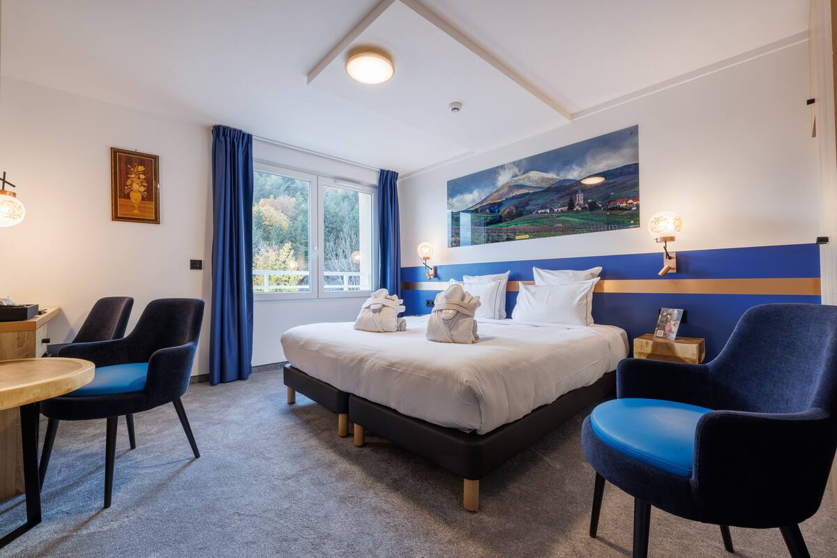 Habitación de hotel con cama, dos sillas, escritorio, ventana con cortinas y un cuadro en la pared.