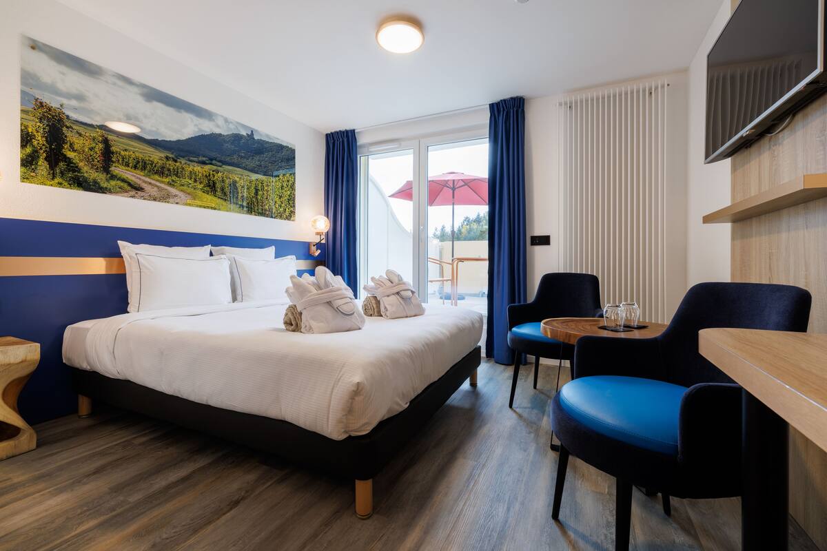 Habitación de hotel con cama, escritorio, silla, televisor y un ventanal con cortinas.