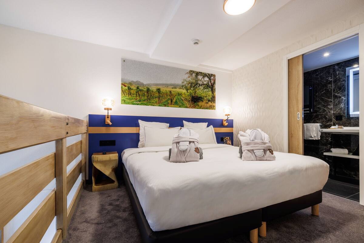 Habitación de hotel con una cama grande, cabecera con diseño azul y dorado, un cuadro de un paisaje en la pared y un baño visible a través de una puerta abierta.