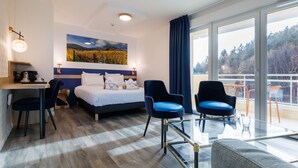 Habitación de hotel con cama, dos sillas, escritorio, una mesa de vidrio y un ventanal con vistas a árboles.