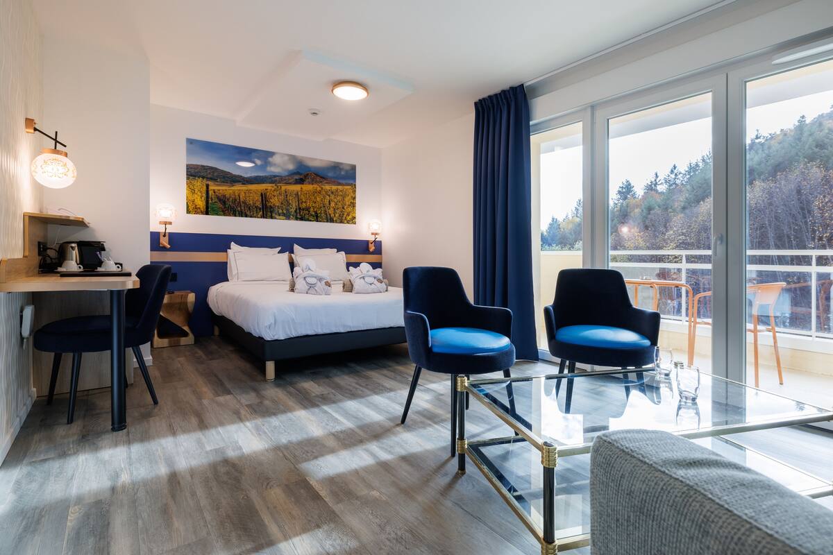 Habitación de hotel con cama, dos sillas, escritorio, una mesa de vidrio y un ventanal con vistas a árboles.