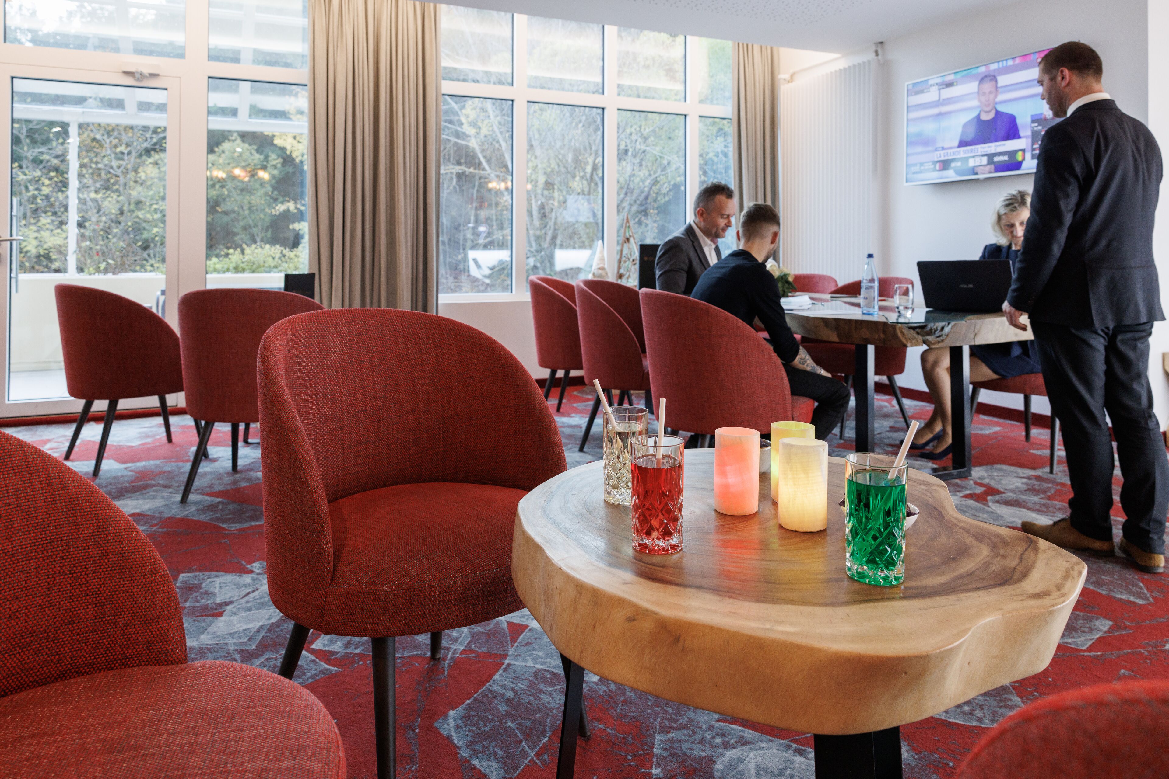 Un moderno vestíbulo de hotel con sillones tapizados en rojo, una mesa de madera con bebidas, ventanas amplias y una pantalla de televisión.