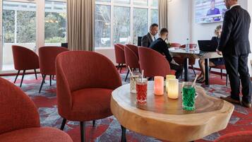 A modern hotel lobby with red upholstered chairs, a wooden table with drinks, large windows, and a TV screen.