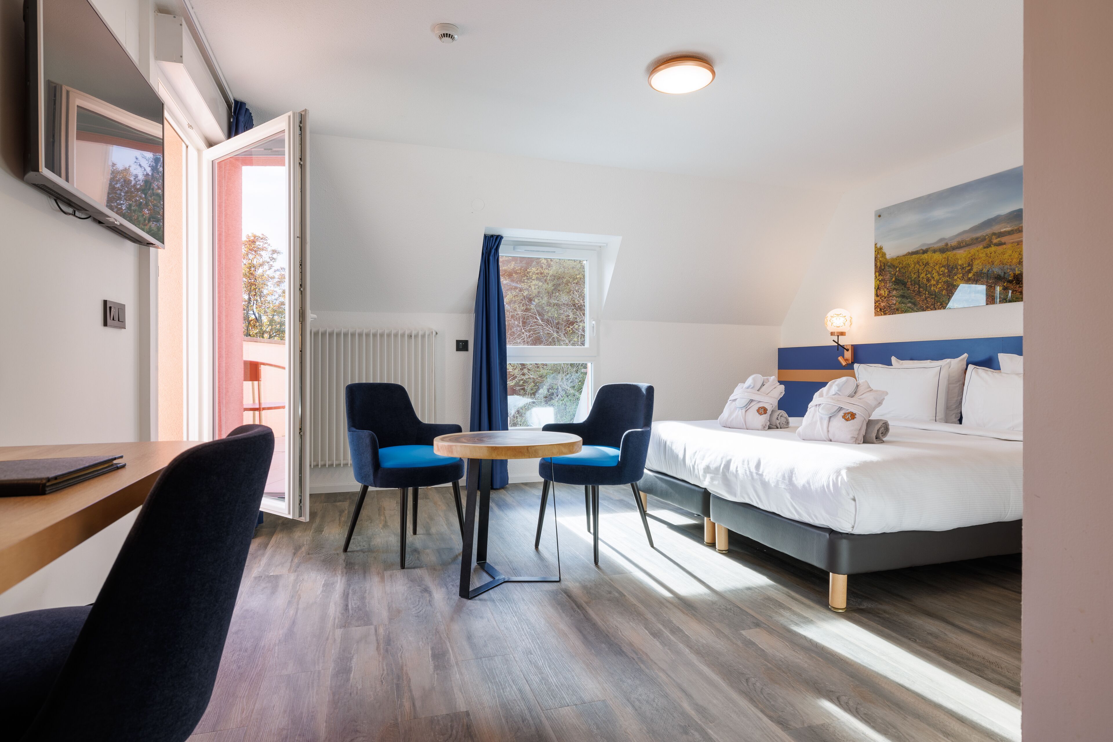 Quarto de hotel com cama, mesa, duas cadeiras, janela com cortinas e um quadro na parede.