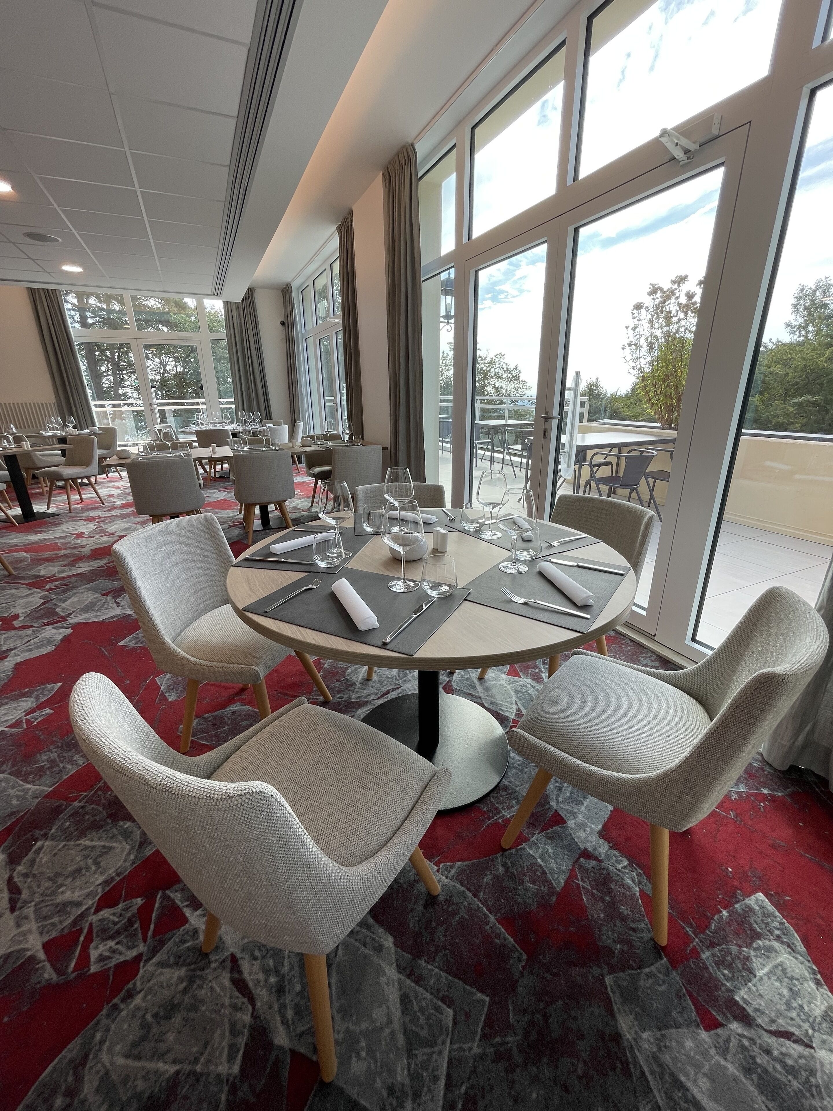 Un espace repas avec des tables rondes, des chaises à revêtement gris et un tapis rouge à motifs.