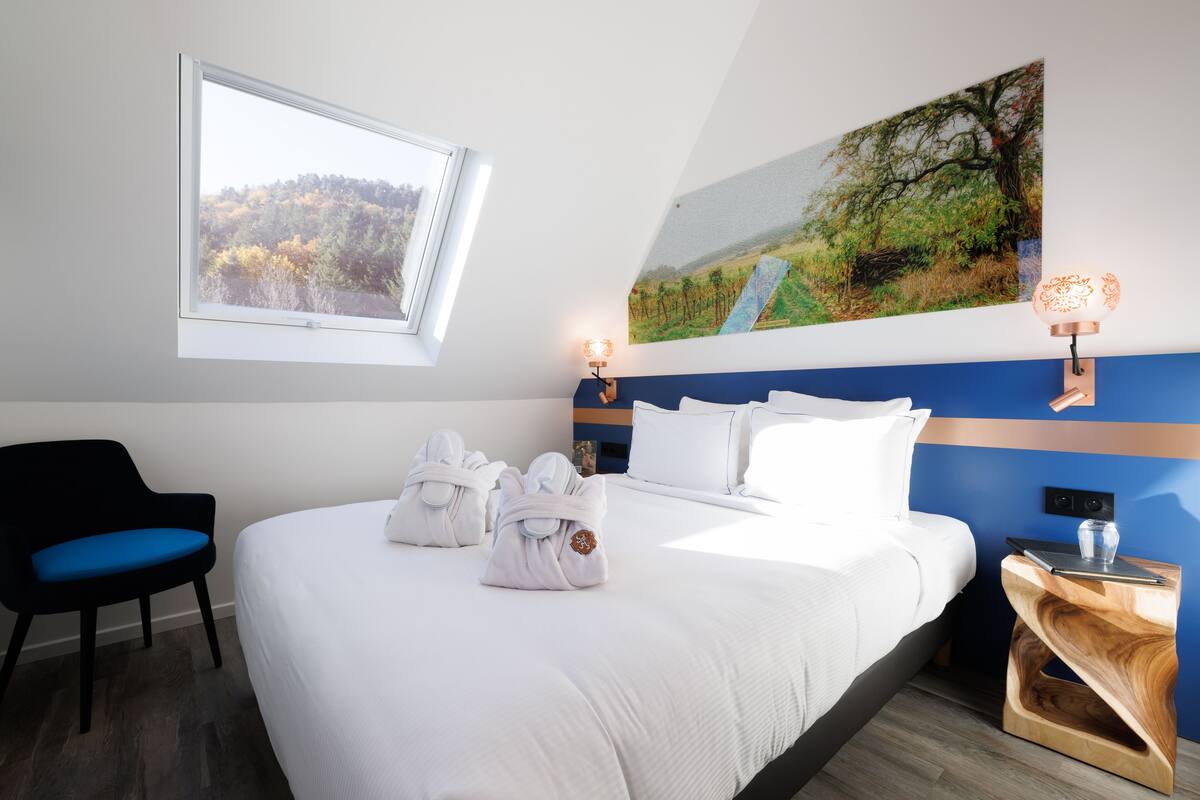 Una habitación de hotel con una cama, dos lámparas de pared, un cuadro, una silla y una mesita de noche con un vaso.
