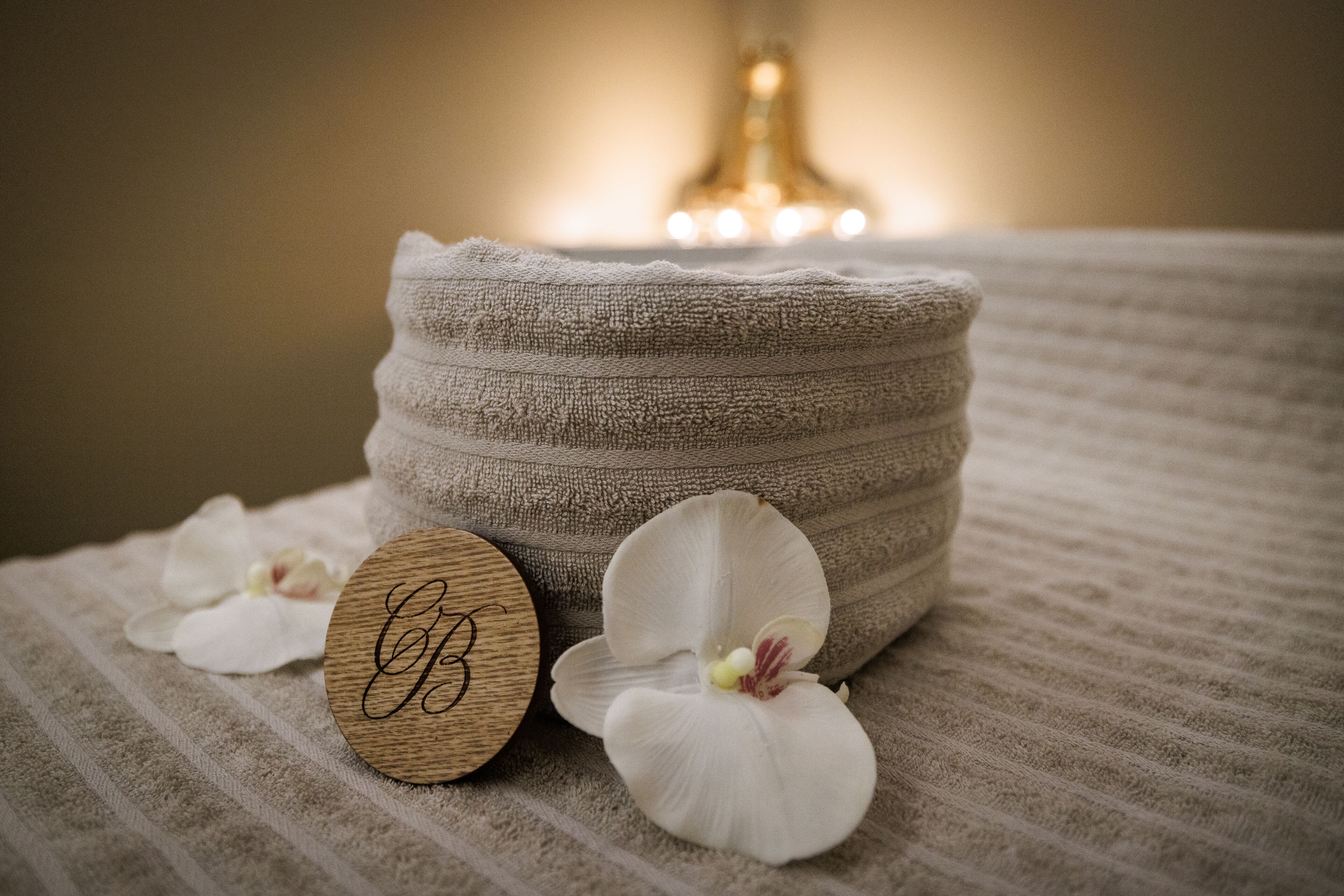 Una toalla beige con textura, una placa de madera con un monograma y pétalos de orquídea blanca sobre una superficie con estampado a rayas.