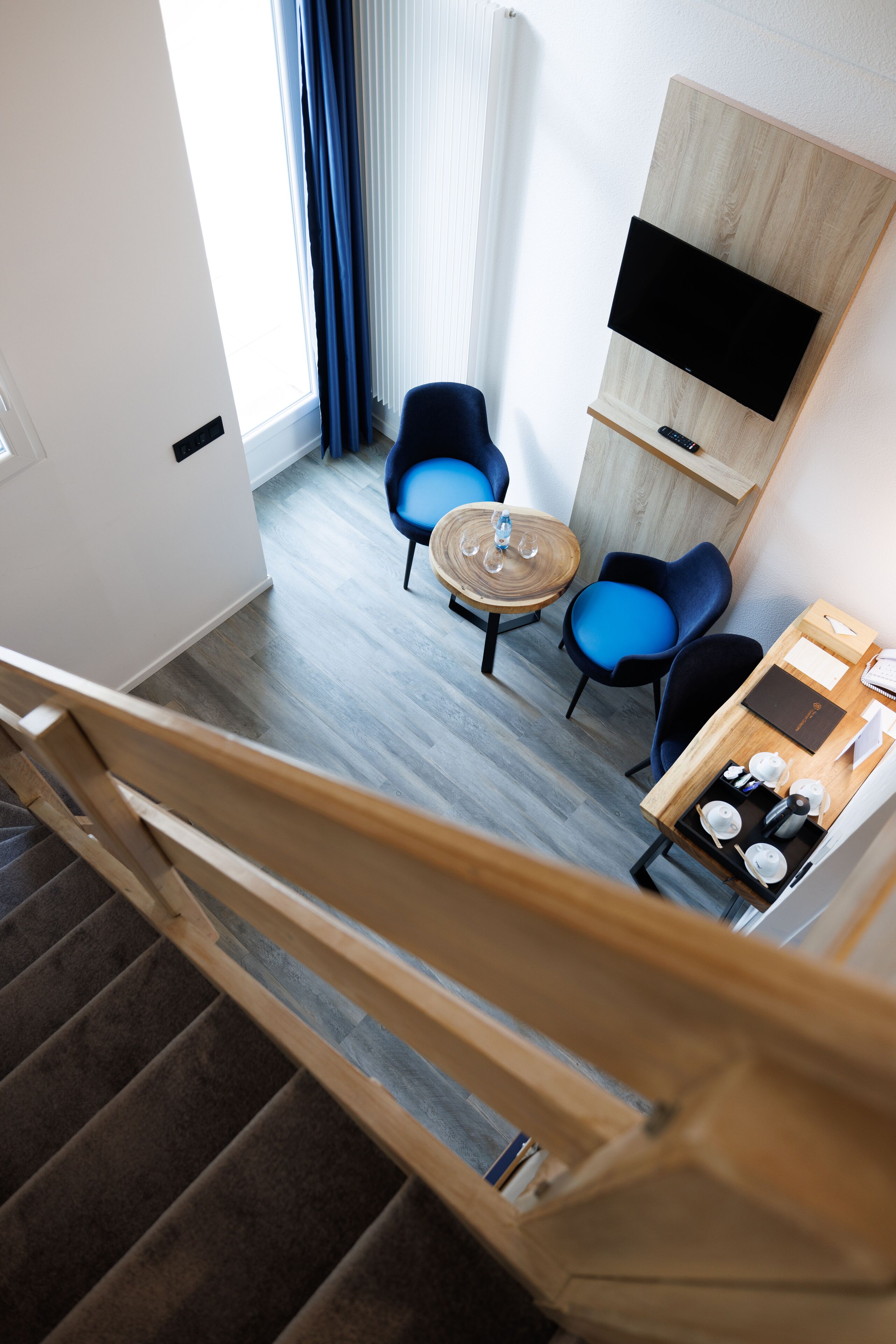 Une chambre d’hôtel moderne avec un escalier en bois, une table ronde entourée de chaises, un téléviseur à écran plat fixé sur un panneau en bois, et une petite table sur laquelle se trouve un plateau avec des tasses et un carnet.