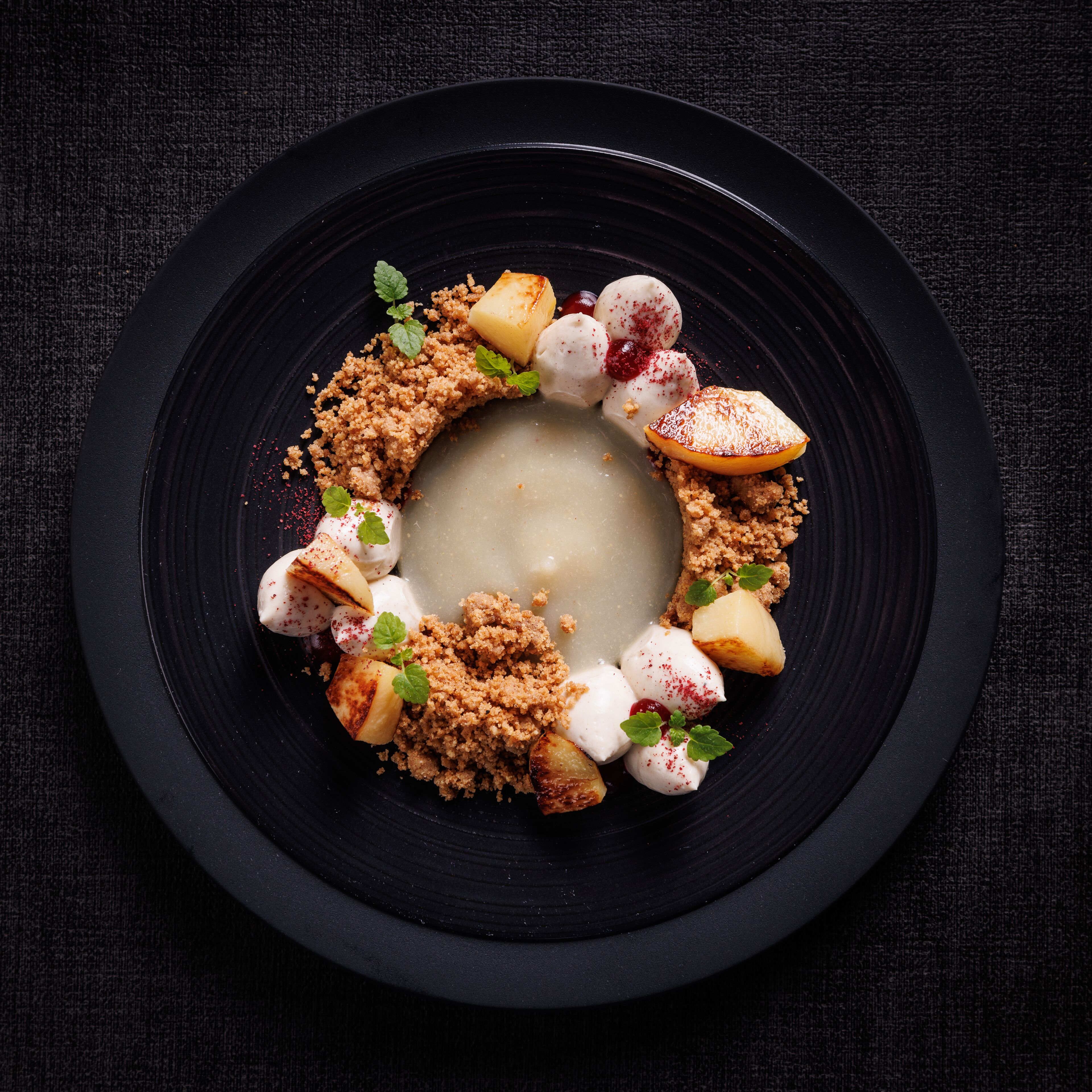 Dessert auf einem schwarzen Teller mit Streuseln, Früchten und Minzblättern.