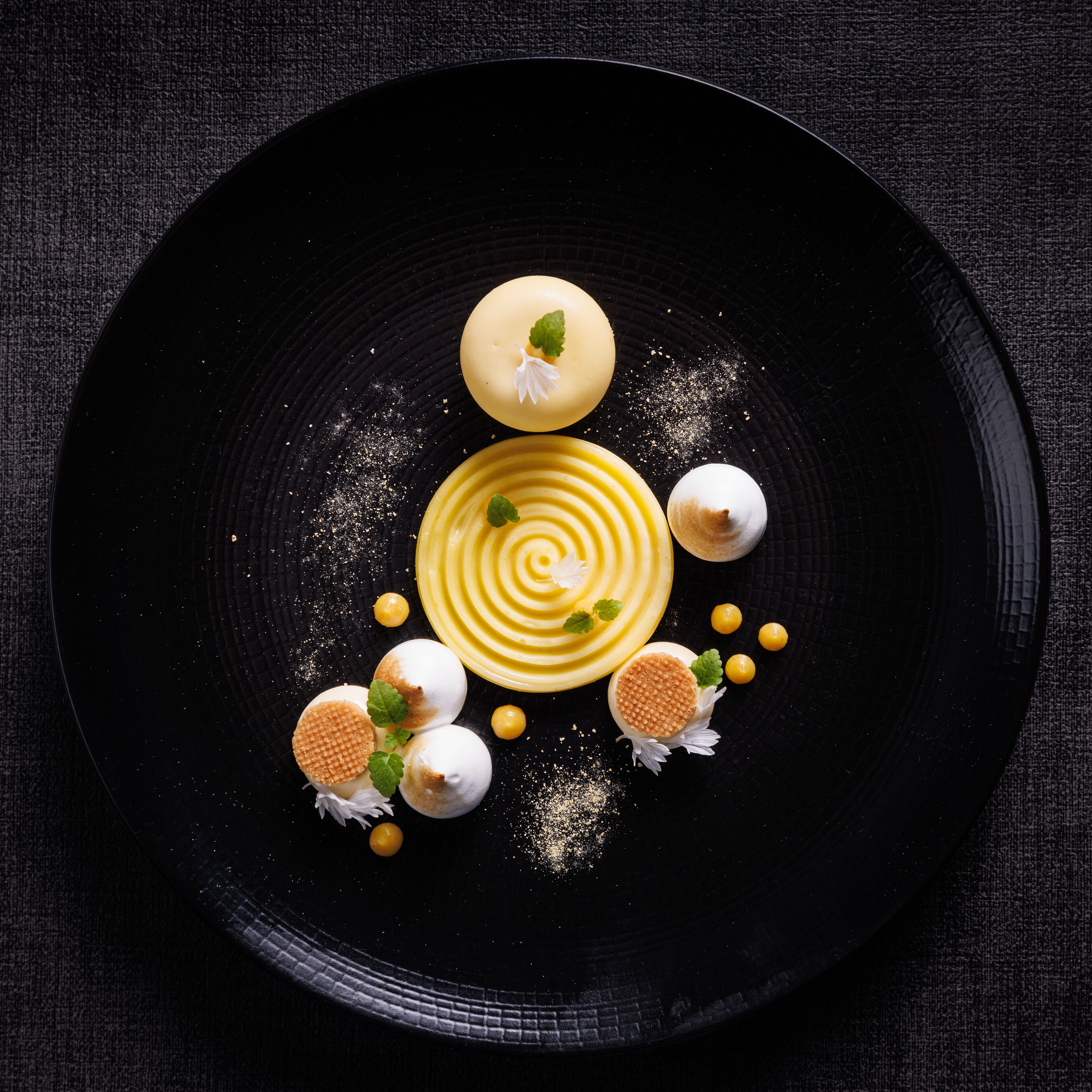 A dessert plate with a central swirl design, surrounded by spherical and waffle-textured items, and small yellow spheres on a dark plate.