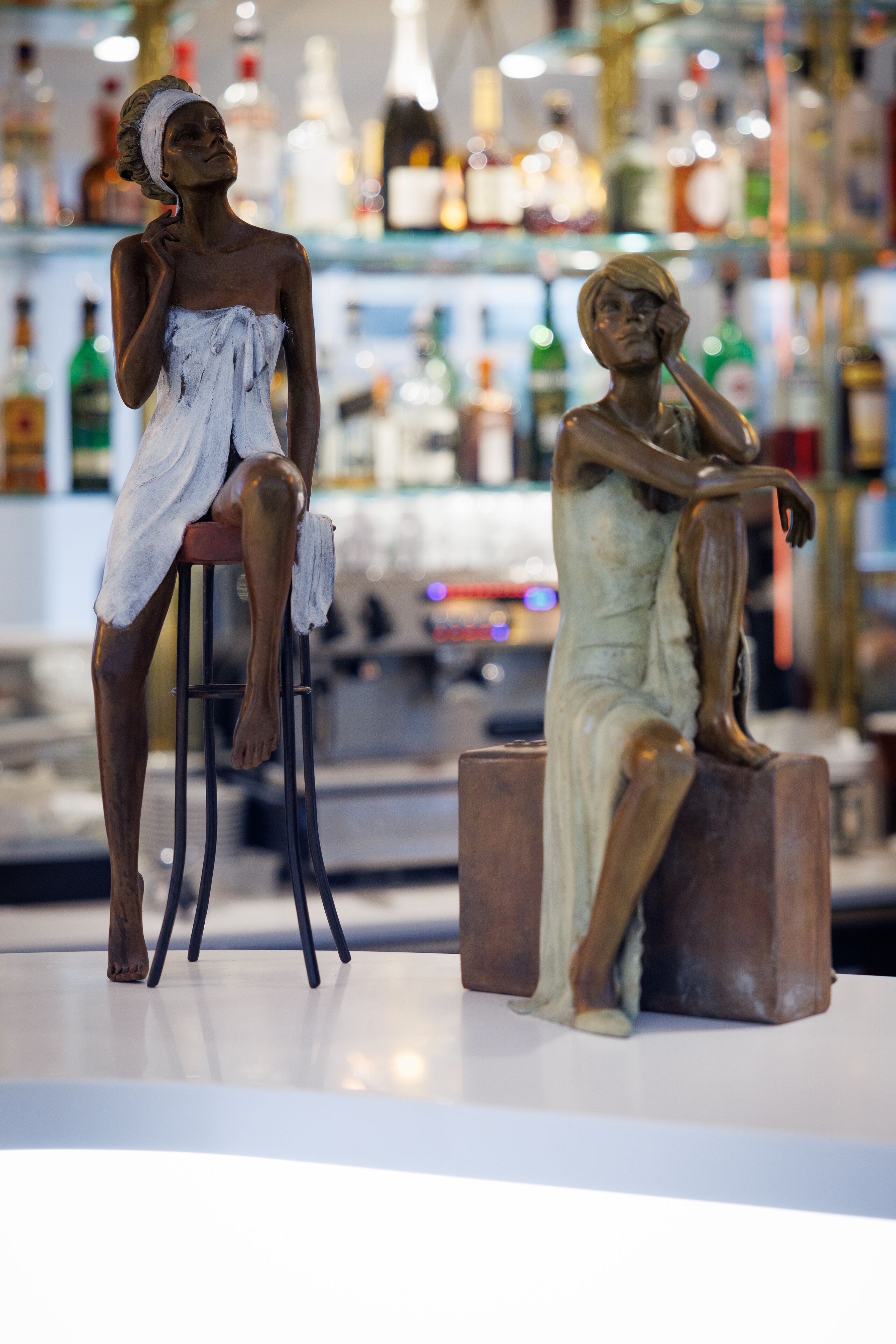 Two bronze statues of seated figures in a bar setting with shelves of bottles in the background.