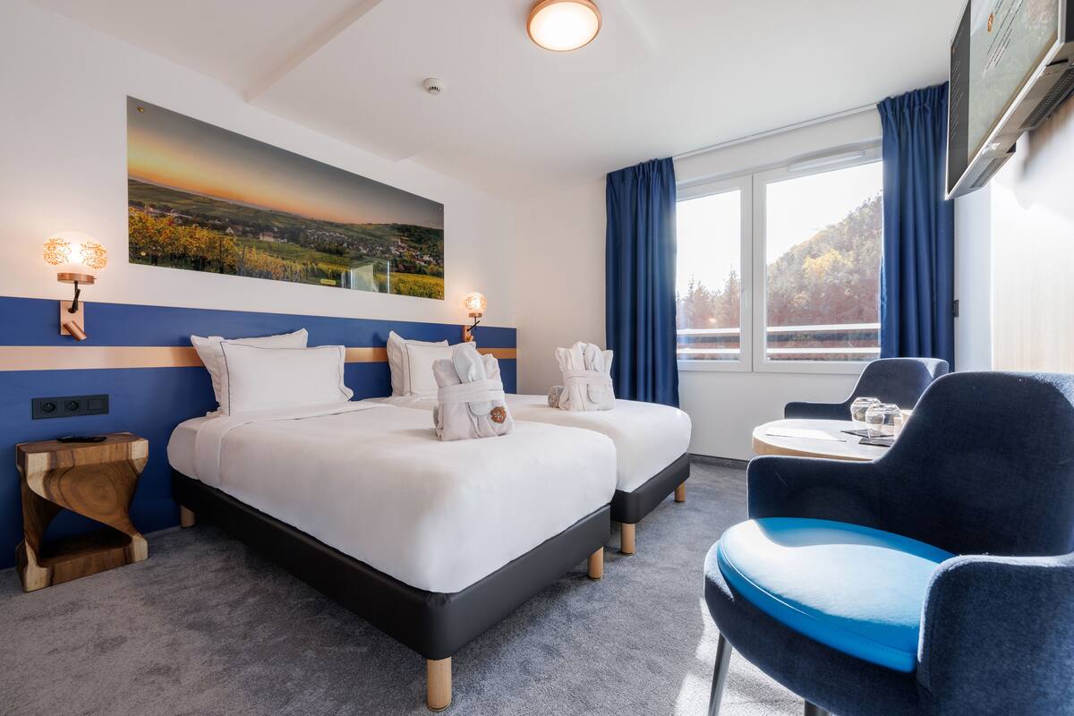 Habitación de hotel con una cama grande, dos mesitas de noche, una silla, una mesita pequeña y una ventana con cortinas.