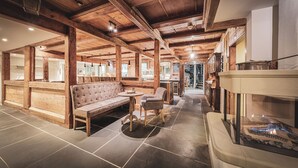 Un intérieur rustique avec des poutres en bois apparentes, un sol carrelé en pierre, un canapé en cuir, une petite table ronde et une cheminée en verre.