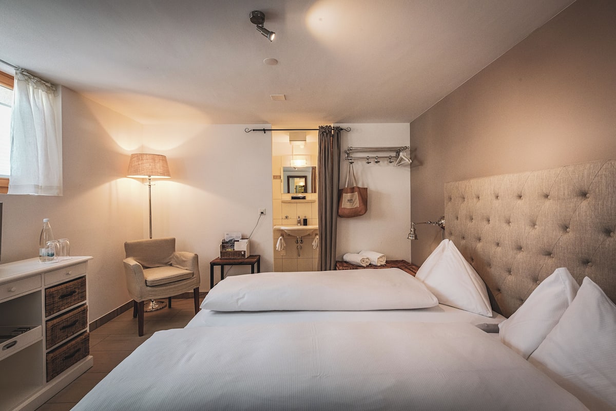 Habitación de hotel con una cama grande, una silla, un escritorio con cajones, una lámpara y un baño a la vista a través de una puerta abierta.