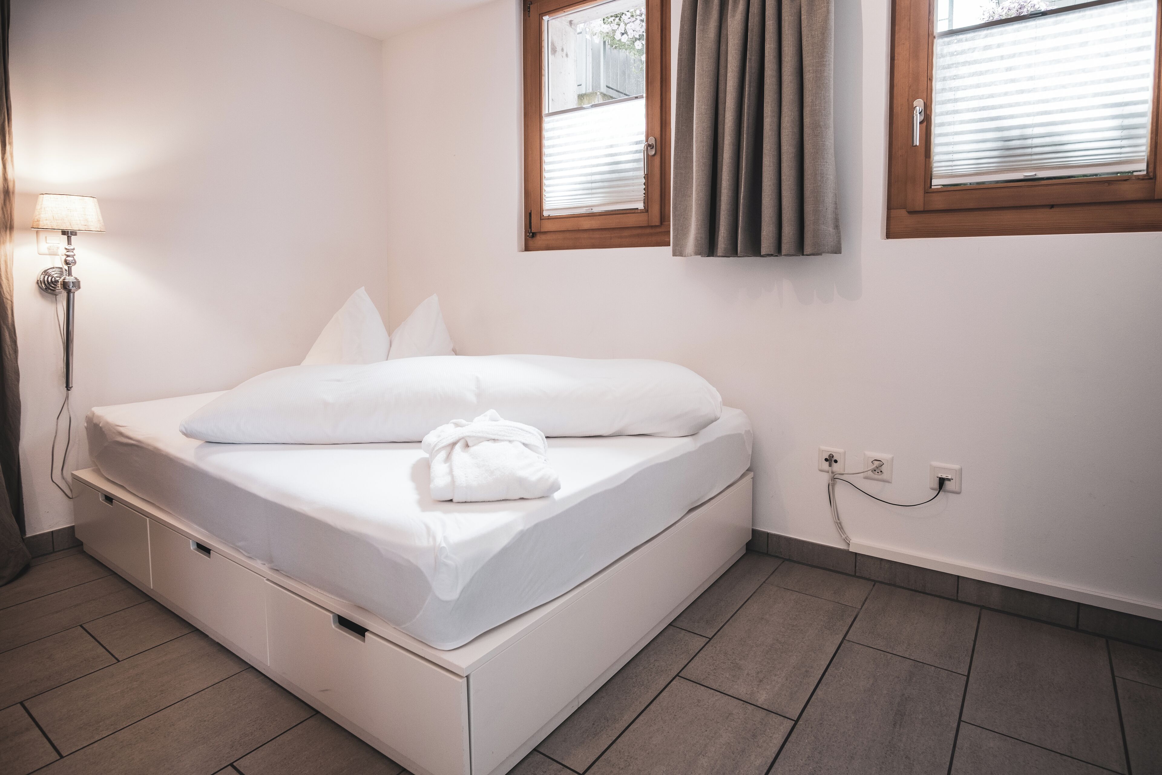Ein ordentlich bezogenes Bett mit weißer Decke und Kissen, eine Nachttischlampe und eine Steckdose an der Wand.