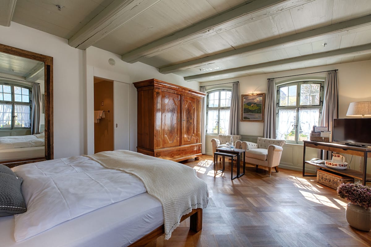 Una habitación amplia con una cama grande, un armario de madera, una mesita, una silla y un televisor.