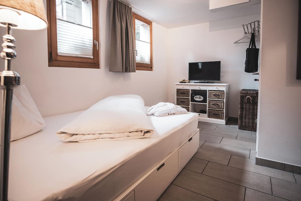 Una cama bien hecha con un colchón y almohadas blancos, un televisor sobre un mueble blanco y una cesta de mimbre en el suelo.