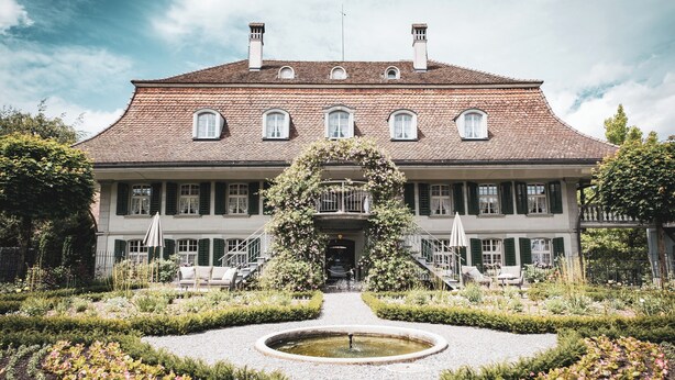 Un grand bâtiment historique à la façade symétrique, doté d’une entrée centrale et d’un jardin à la française agrémenté d’une fontaine.