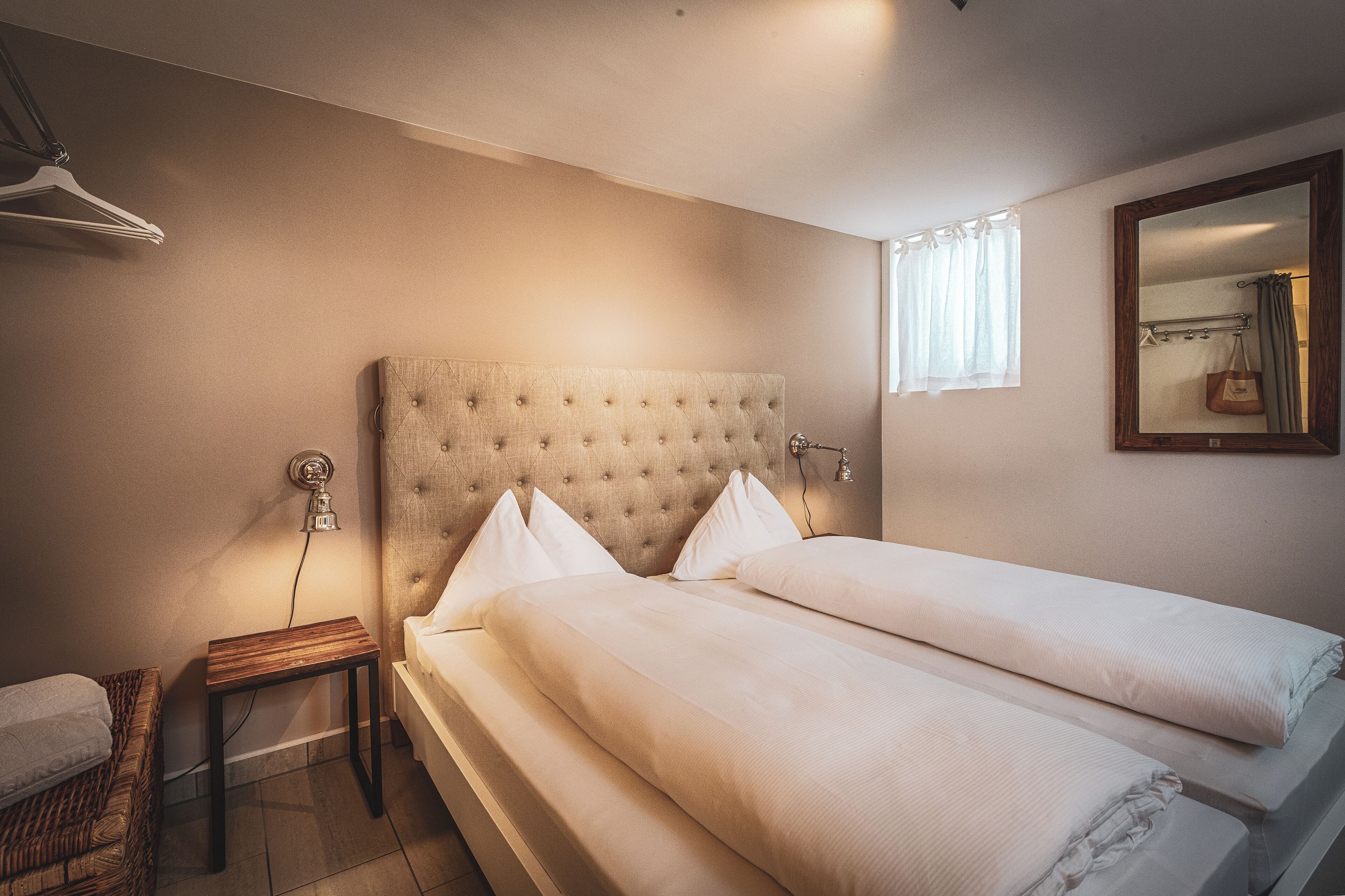 Une chambre avec deux lits, une table de chevet en bois, une lampe suspendue, un miroir et une fenêtre avec des rideaux.