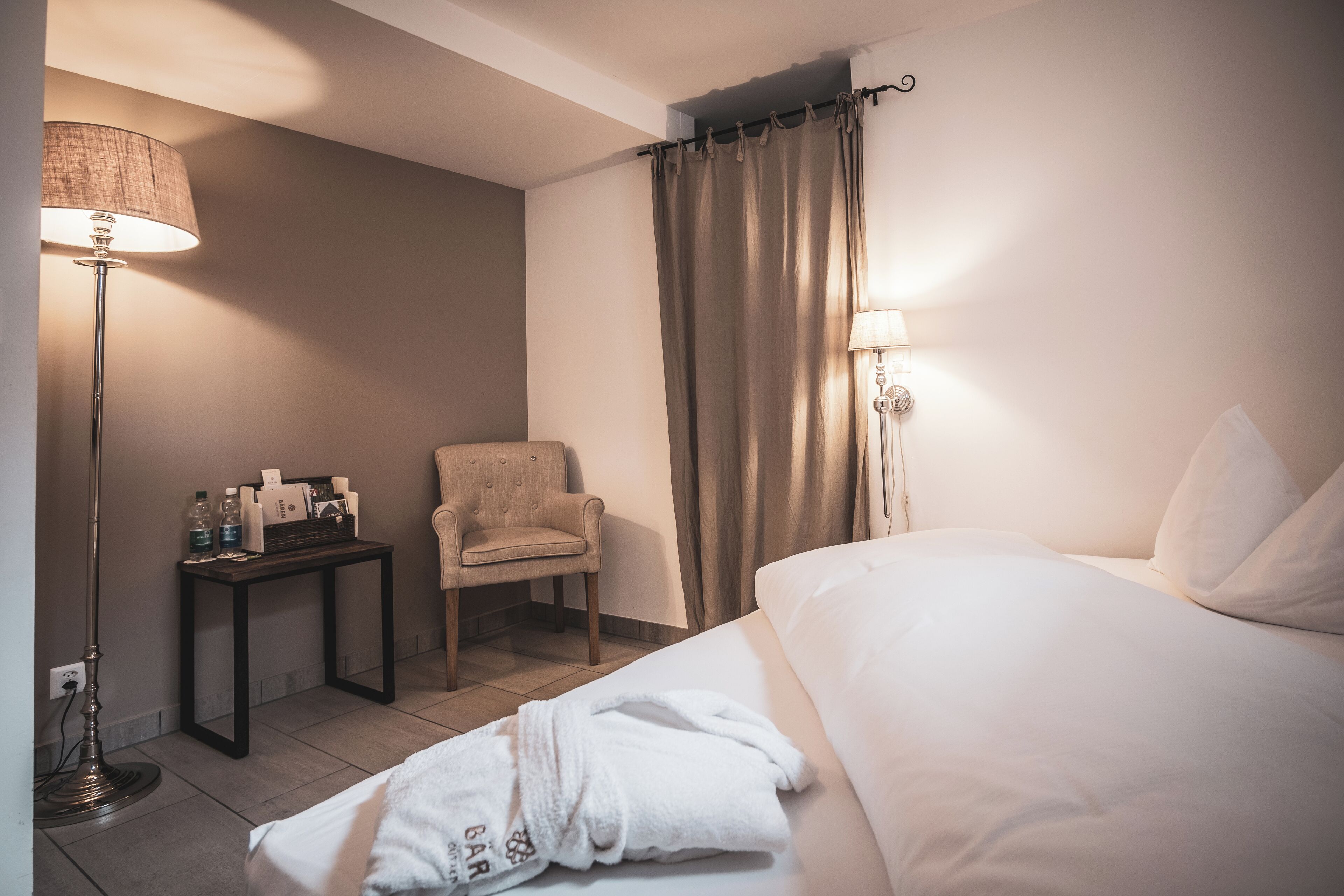 Een hotelkamer met een bed, een stoel, een tafel met toiletartikelen, een lamp en een gordijn.