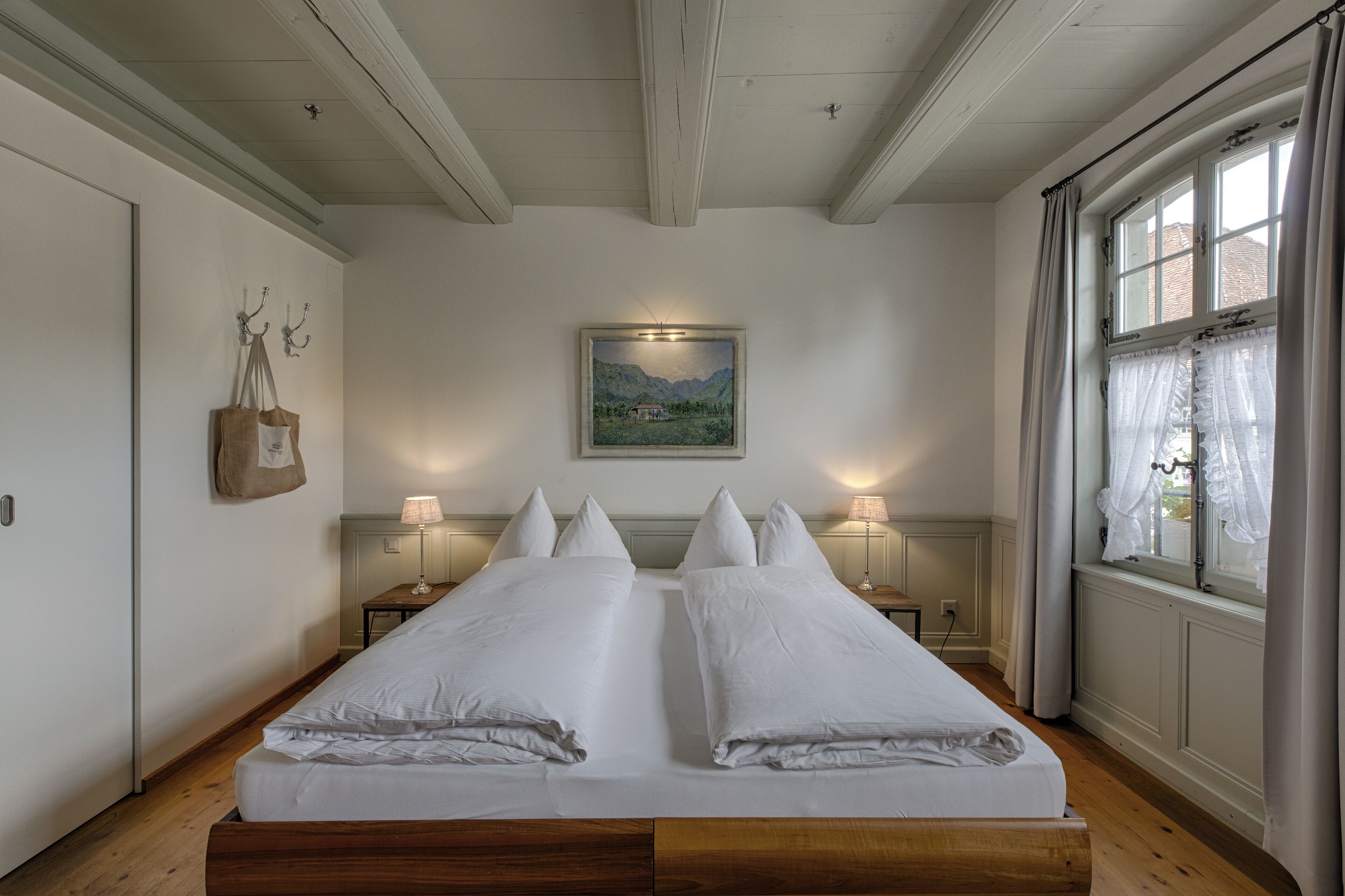 A double bed with white linens, two bedside tables with lamps, a painting on the wall, and a window with curtains.